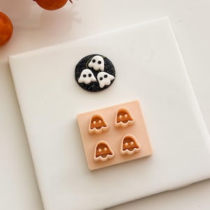 Puede incluir: Un disco circular negro brillante con tres pequeñas figuras de fantasmas blancos con ojos negros, colocado sobre un molde cuadrado de color melocotón que contiene cuatro recortes con forma de fantasma y dos pequeñas hendiduras oculares.