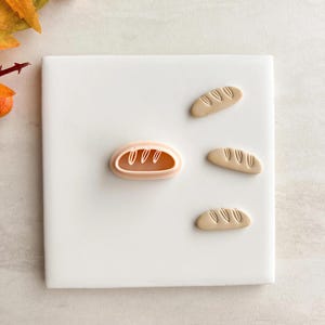 May include: A light pink bread-shaped cookie cutter with three beige bread-shaped cookies. The cookie cutter is in the shape of a baguette. The cookies have detailed lines on top. The items are on a white surface.