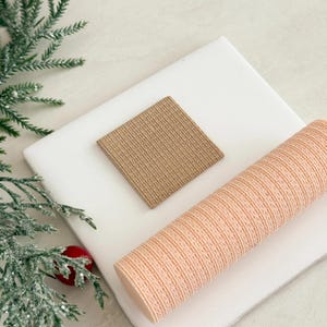 May include: A square, tan coaster and a rolled, peach-colored item with a textured surface are displayed on a white, square surface. A sprig of evergreen with a red berry is in the background.