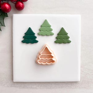 May include: A selection of Christmas tree-shaped cookie cutters and cookies in shades of green and peach. The cookies are arranged on a white surface, with red berries and green leaves in the top left corner.