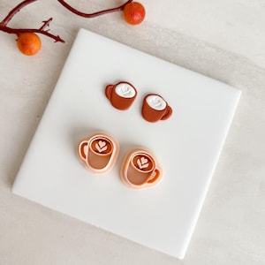 May include: Four coffee-themed items are displayed on a white surface. Two small, brown, cup-shaped earrings with white foam designs are at the top. Below are two coffee cup-shaped cookie cutters with a heart design.