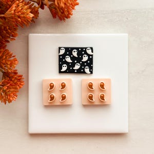May include: A rectangular black plaque featuring a white ghost and polka dot pattern, accompanied by two light peach square plaques, each displaying four small, brown, teardrop-shaped ghost figures with white outlines and black eyes.