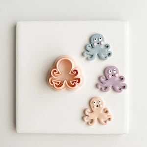May include: A collection of octopus-shaped items. There is a light peach-colored cookie cutter and three small octopus figurines in blue, purple, and tan. All items have a cartoonish design with large eyes and smiling faces.