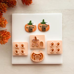 Puede incluir: Dos dijes con forma de casa calabaza con tallos verdes y pequeños fantasmas blancos, junto a cinco cortadores de plástico color melocotón claro. Los cortadores incluyen formas para cuatro fantasmas pequeños, una casa calabaza, un sombrero, una puerta y ventanas.