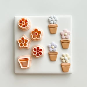 May include: A collection of flower and pot-shaped cookie cutters in various designs and colors. The cutters are in shades of peach and orange, with some featuring red and white accents. The flower pots are light brown, and the flowers are pastel colors.