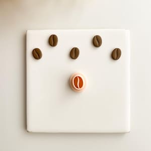 May include: A white square tile with six brown coffee bean shapes. One coffee bean shape is a cookie cutter. The other five coffee bean shapes are scattered across the tile. The background is white.