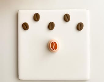 May include: A white square tile with six brown coffee bean shapes. One coffee bean shape is a cookie cutter. The other five coffee bean shapes are scattered across the tile. The background is white.