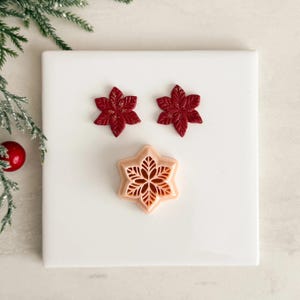 May include: Three Christmas-themed cookie cutters and decorations. Two dark red poinsettia-shaped cutters and one light pink snowflake-shaped cutter are displayed on a white surface. The poinsettias have detailed leaf patterns.