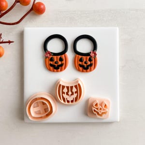 May include: A selection of Halloween-themed cookie cutters. Two orange pumpkin-shaped cutters with black handles and smiling faces are shown, alongside three other cutters in different shapes. The cutters are on a white surface.