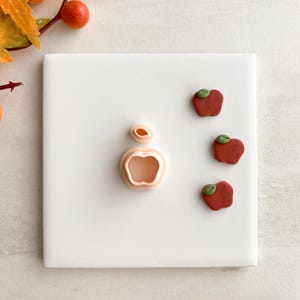 May include: A peach-coloured apple-shaped cookie cutter and a stem cutter. Three small, red apple-shaped biscuits with green leaf accents are arranged on a white square surface. The image has an autumnal theme.
