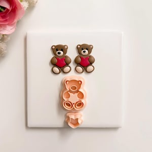 May include: Two brown teddy bear figurines with red hearts, and a pink teddy bear cookie cutter with a heart-shaped cutter. The bears have black eyes and tan paws. The items are on a white surface.