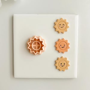 May include: A light peach-colored sun-shaped cookie cutter next to three smiling sun-shaped decorations in shades of orange and yellow. The cookie cutter has a cross-shaped design in the center. The objects are on a white surface.
