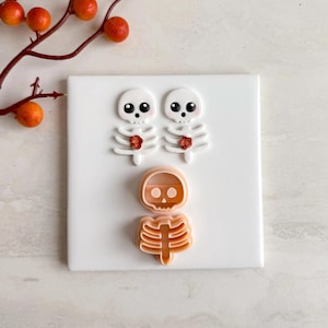 Puede incluir: Se muestran tres cortadores de galletas con temática de esqueleto y dos decoraciones con forma de esqueleto sobre una superficie blanca. Las decoraciones tienen calaveras blancas con ojos negros y detalles de flores rojas. Los cortadores de galletas son de color naranja claro.
