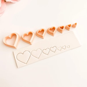 Op de afbeelding: Een set perzikroze hartvormige koekjessnijders in verschillende maten, gerangschikt op een wit oppervlak. De uitstekers liggen naast een stuk deeg met hartafdrukken, ideaal voor bakken of knutselen.
