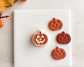 May include: A collection of Halloween-themed pumpkin-shaped cookie cutters in various shades of orange and brown. One cutter is light orange, and the other three are darker shades of orange and brown. The cutters are arranged on a white square surface.