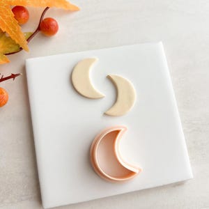 Op de afbeelding: Drie halvemaanvormige items. Twee zijn lichtbeige, mogelijk koekjes of fondant uitsteekvormen. De derde is een roze plastic koekjessteker. De items liggen op een wit oppervlak, met herfstbladeren op de achtergrond.