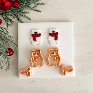 May include: A selection of bear-shaped cookie cutters and fondant decorations. The fondant bears are white with red scarves, and the cookie cutters are light brown. A sprig of evergreen and red baubles are in the background.