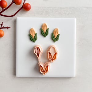 May include: A selection of corn-shaped cookie cutters and corn decorations. The cutters are pale pink, and the corn decorations are yellow with green husks. The items are arranged on a white surface, with a branch of orange berries in the background.