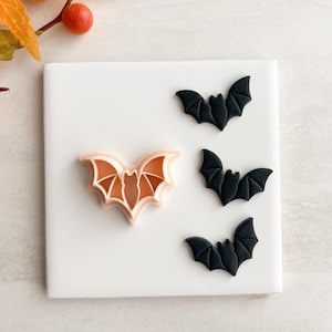 Puede incluir: Una baldosa cuadrada blanca con un cortador de galletas en forma de murciélago y tres galletas negras en forma de murciélago. El cortador de galletas es de color naranja claro y las galletas son negras. La imagen tiene un tema de Halloween.