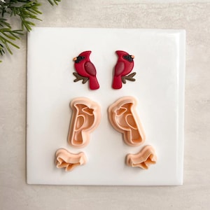May include: A collection of red cardinal bird-shaped cookie cutters and matching cookie dough cutouts, arranged on a white surface. The cardinals are perched on brown branches. The cookie cutters and cutouts are in a light peach color.