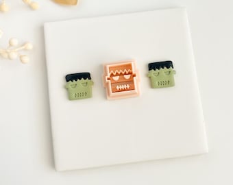 May include: Three Halloween-themed decorative magnets. Two green magnets with black hair and a stitched mouth design flank a light orange magnet with a Frankenstein monster face. The magnets are on a white surface.