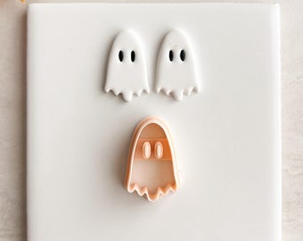 May include: Three ghost-shaped cookie cutters. Two white ghosts with black eyes are above a peach-colored cutter. The cutters are on a white surface, with a sprig of berries and leaves in the background. Halloween-themed.