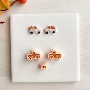 May include: A collection of fall-themed craft supplies. Two pairs of beige car-shaped earrings with pumpkin accents and black wheels. Below are three car-shaped cookie cutters in a light orange color, also with pumpkin accents. The items are on a white surface.