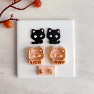May include: Two black cat-shaped figurines and two cat-shaped cookie cutters with striped details are displayed. A small, rectangular piece with two triangle shapes is also present. The items are arranged on a white surface.