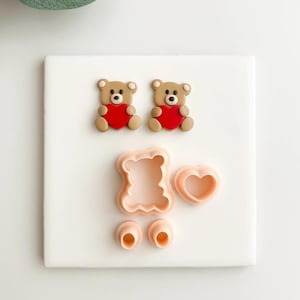 May include: Two small brown teddy bear figurines with red heart accents. Also shown are the components of a teddy bear cookie cutter set, including a bear outline, heart, and eye shapes. The items are arranged on a white surface.