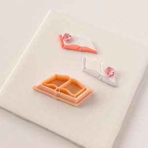 May include: An assortment of book-shaped items. There is a peach-colored cookie cutter in the shape of an open book. Two open book-shaped decorations are also present, one with a peach-colored cover and the other with a white cover, each adorned with a pink flower.