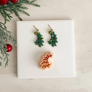 Op de afbeelding: Een paar kerstboomoorbellen met goudkleurige haken en een bijpassende koekjesvorm. De oorbellen zijn groen met kleurrijke ornamenten. De koekjesvorm is perzik van kleur en heeft de vorm van een kerstboom.