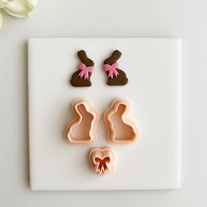 Puede incluir: Una selección de cortadores de galletas con temática de Pascua. Dos galletas marrones con forma de conejito con lazos rosas se muestran encima de tres cortadores de galletas rosas: dos contornos de conejito y un lazo. Los artículos están dispuestos sobre una superficie blanca.