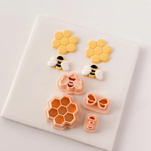 May include: A collection of yellow and peach-colored cookie cutters and fondant molds in a bee and honeycomb theme. Includes two honeycomb shapes, two bees, two bow shapes, a large honeycomb shape, a bow shape, and a small shape.