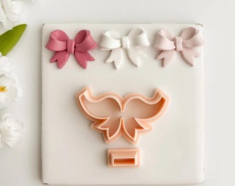 May include: A selection of bow-shaped fondant decorations in shades of pink and white, displayed on a white surface. A bow-shaped cookie cutter is also present, indicating a baking or crafting theme. The bows are in various shades of pink.
