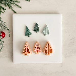 Könnte beinhalten: Sechs Ausstecher in Form von Weihnachtsbäumen in verschiedenen Designs und Farben. Die Ausstecher sind in Grün- und Pfirsichtönen gehalten und auf einer weißen Oberfläche angeordnet. Das Bild enthält auch einen Tannenzweig und eine rote Dekoration.