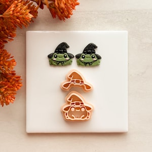 May include: Two small green frog figures wearing black glittery witch hats with tiny black bows, positioned above two light peach-colored plastic cookie cutters, one shaped like a witch hat and the other shaped like a frog wearing a witch hat.