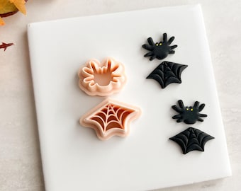 May include: Halloween-themed cookie cutters and decorations. The image features a spider, a spiderweb, and a bat cookie cutter, along with two black bat and spider decorations. The cutters are light pink, and the decorations are black.
