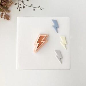 May include: A collection of lightning bolt-shaped objects in various pastel colors. The largest is a light orange color with a pink outline. Other bolts are light blue, pale yellow, and gray. The objects are arranged on a white square surface.