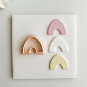 May include: A light peach-colored polymer clay cutter in the shape of an arch, with three arch-shaped clay pieces in pink, white, and yellow. The items are arranged on a white square surface, with a white flower in the upper left corner.