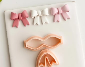 May include: Three decorative bows in shades of pink and white are arranged on a white surface. Below, three peach-coloured cookie cutters in bow shapes are displayed. The image showcases items for crafting or baking, with a focus on bows.