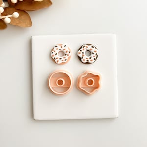 May include: A square white surface displays two miniature donut-shaped food items with colorful sprinkles and two pink donut-shaped cookie cutters. One donut has a white base with orange and brown sprinkles, and the other has a brown base with sprinkles.