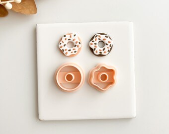 May include: A square white surface displays two miniature donut-shaped food items with colorful sprinkles and two pink donut-shaped cookie cutters. One donut has a white base with orange and brown sprinkles, and the other has a brown base with sprinkles.