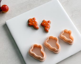 May include: Two orange cookies with white dots and brown edges, alongside three peach-colored cookie cutters. The cookies are shaped like a mushroom. The cutters are also mushroom-shaped, perfect for baking.