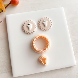 May include: A collection of Halloween-themed cookie cutters. Two round cutters with scalloped edges and a ghost design, a round ring cutter, and a ghost-shaped cutter. The cutters are peach-colored and sit on a white surface.