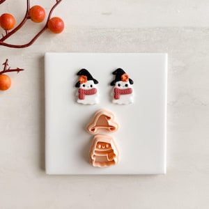 May include: Halloween-themed cookie cutters and decorated biscuits. Two ghost-shaped biscuits with black witch hats, red scarves, and orange flower accents. A witch hat and ghost biscuit cutter are also present. The items are on a white surface.