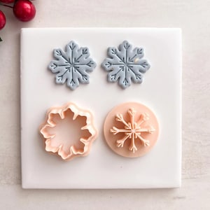 May include: Four snowflake-shaped items on a white surface. Two are light blue, detailed snowflakes. One is a peach-coloured cookie cutter. The last is a peach-coloured snowflake stamp.