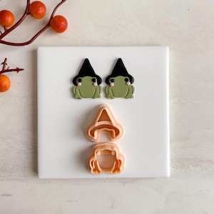 May include: Two green frog-shaped earrings wearing black witch hats, and two cookie cutters: a witch hat and a witch's face. The earrings and cookie cutters are on a white surface, with orange berries in the background. The earrings are a Halloween theme.