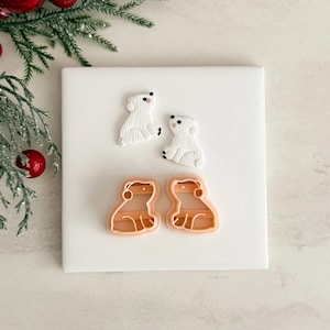 May include: Two white polar bear figurines with black details and two light orange bear-shaped cookie cutters are displayed on a white surface. The figurines have textured fur details. The cookie cutters have a smooth, rounded design.