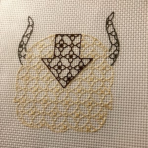 May include: A cross-stitch embroidery design of a stylized animal with a brown arrow-shaped head and two curled horns. The animal's body is filled with a pattern of small, gold circles.