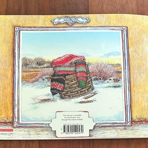 May include: The back cover of a children's book shows a winter scene with a woven basket filled with colourful blankets and scarves. The illustration is framed with a wooden border and a decorative element at the top.